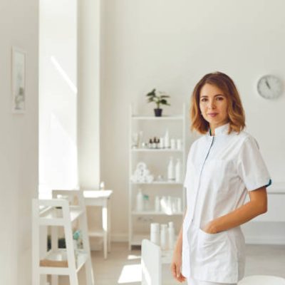 Young smiling woman cosmetologist or dermatologist standing and looking at camera in light beauty spa salon over cosmetics at background. Cosmetology and skincare concept